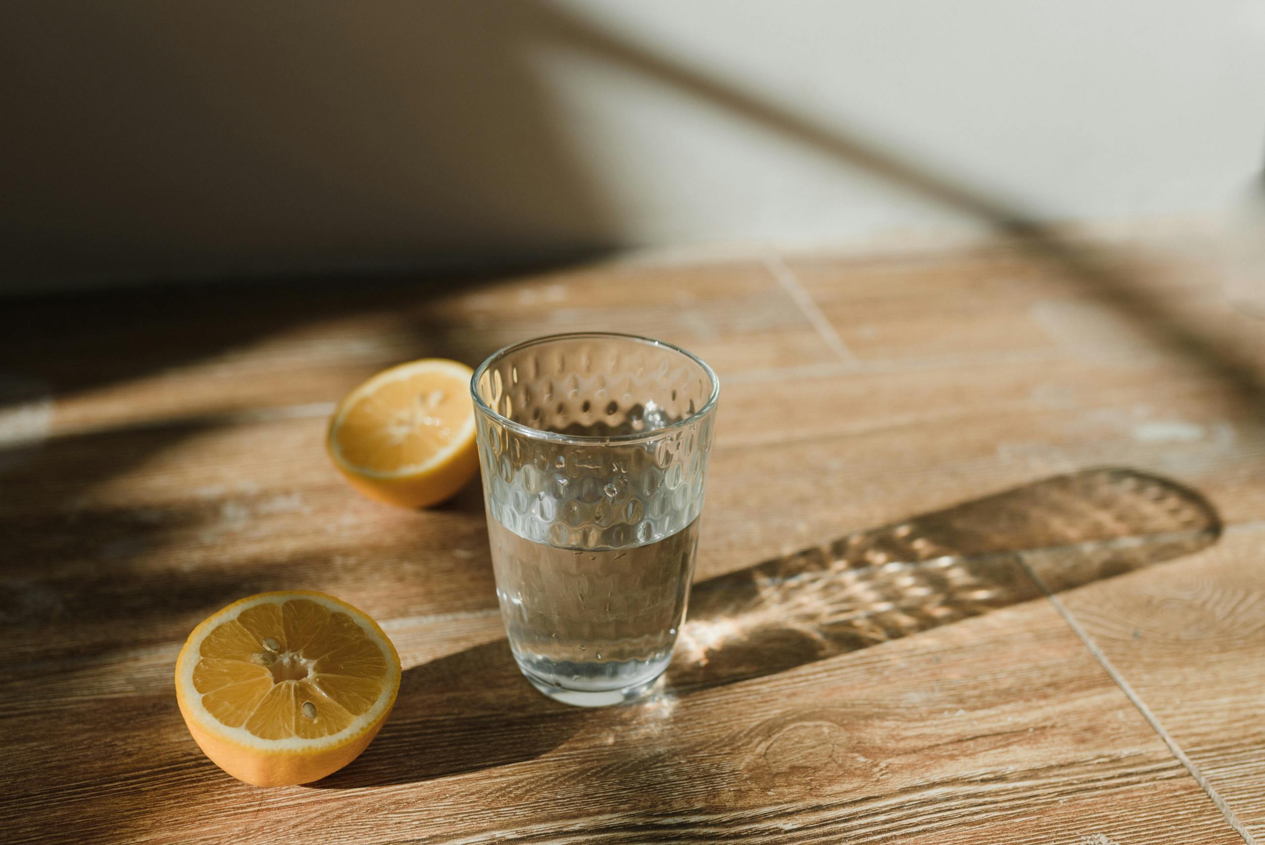 Clear glass of water with sliced lemons on a wooden surface in natural sunlight.
